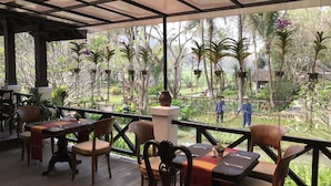 An outdoor dining area with wooden tables and chairs, a covered roof, and a view of a garden with various plants and a pond. - Santi Resort & Spa (Luang Prabang)