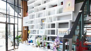 A modern interior with a white shelving unit displaying various items, a reception desk with a vase of flowers, and a staircase leading to an upper level. - The Bed Hatyai (Hat Yai)