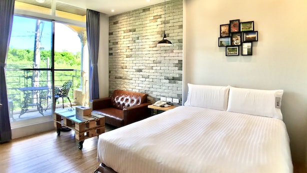 A hotel room with a large bed, a leather armchair, a small table with a phone, and a view of greenery through a window. - Martin Wales Hotel Kenting (Hengchun)