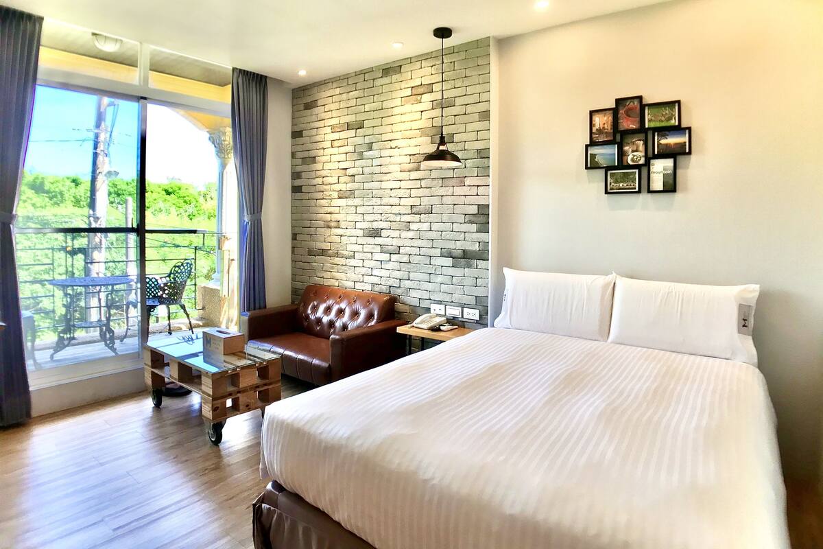 A hotel room with a large bed, a leather armchair, a small table with a phone, and a view of greenery through a window.