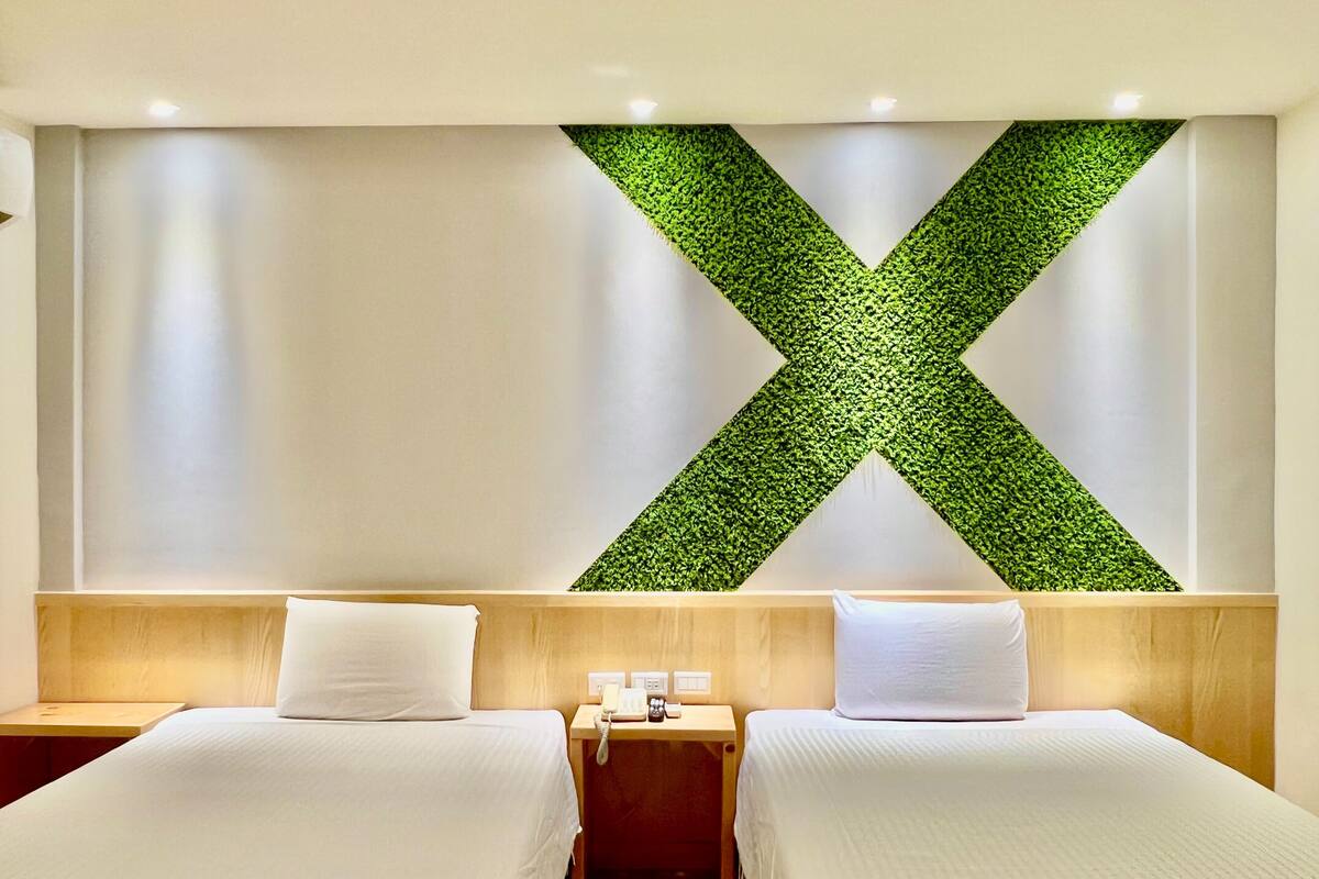 A hotel room with two beds, a wooden headboard, a green wall accent, and a small bedside table with a phone.