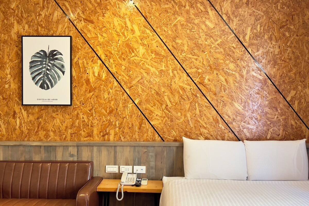 A hotel room with a wooden wall, a framed leaf artwork, a brown leather sofa, a wooden nightstand with a phone, and a bed with white linens.