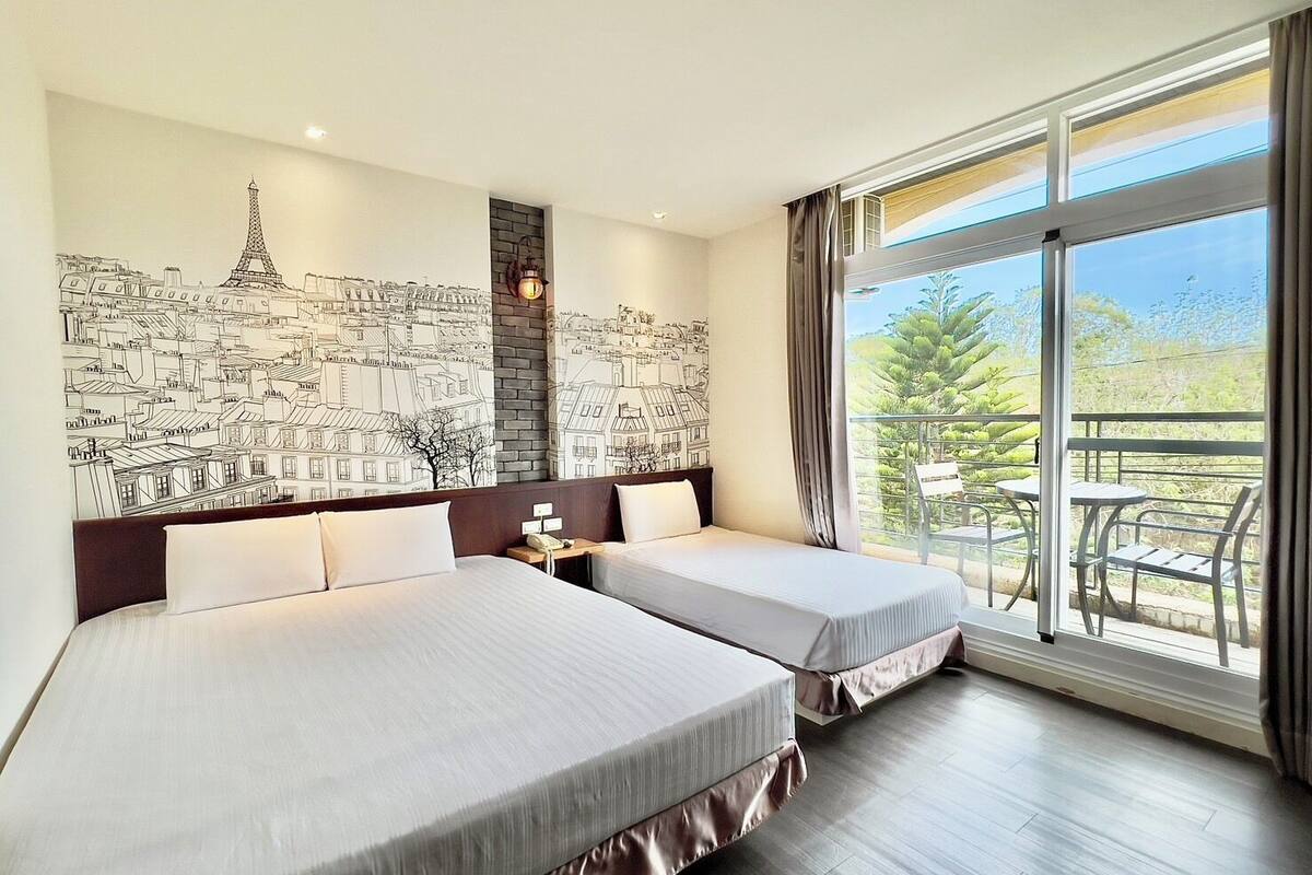 A hotel room with two beds, a cityscape mural, a balcony with outdoor seating, and a view of greenery.