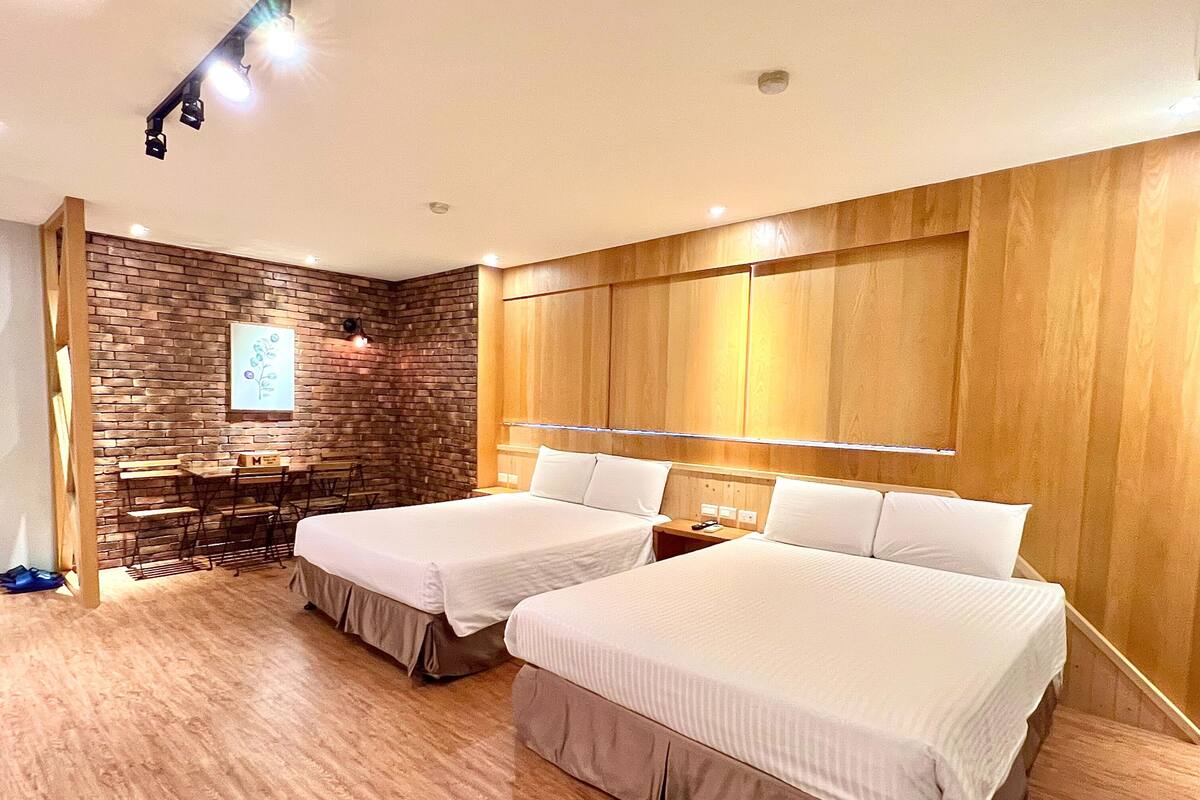 A hotel room with two beds, a brick accent wall, a wooden panel wall, and a small desk with a chair.
