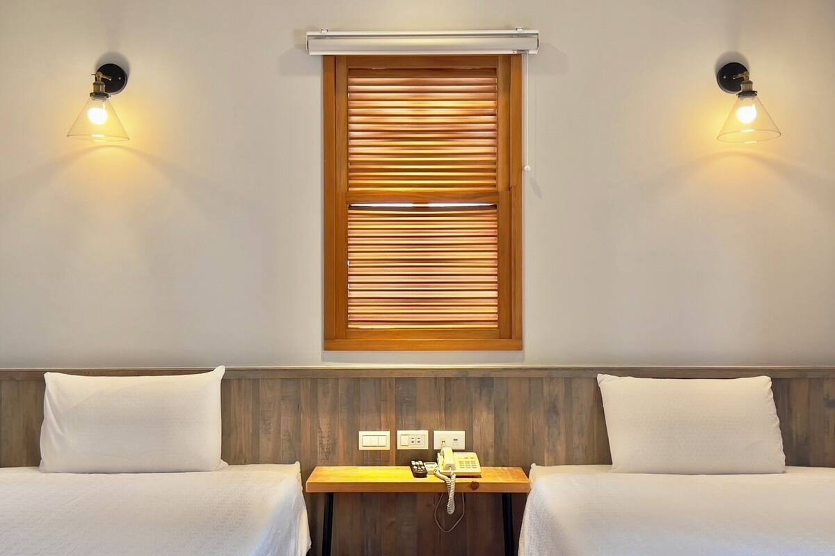 A hotel room with two beds, a wooden headboard, a small wooden table with a telephone, and a window with blinds.
