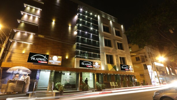 Un hôtel à plusieurs étages, doté d’une enseigne lumineuse indiquant les noms « Prajwal » et « mango hotels ».