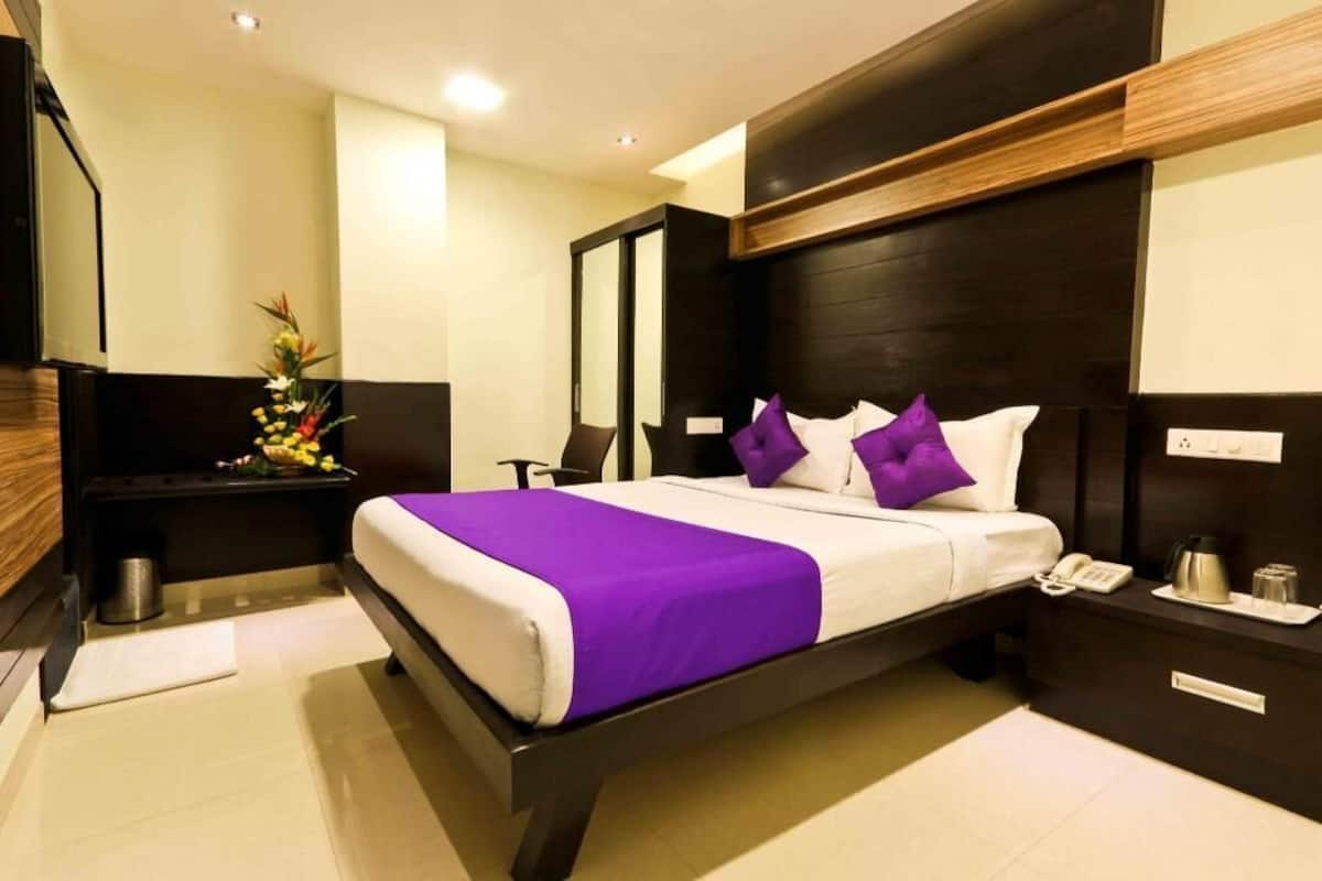 A modern hotel room with a large bed, a nightstand with a phone and a coffee maker, a small table with a vase of flowers, and a dark wooden headboard.