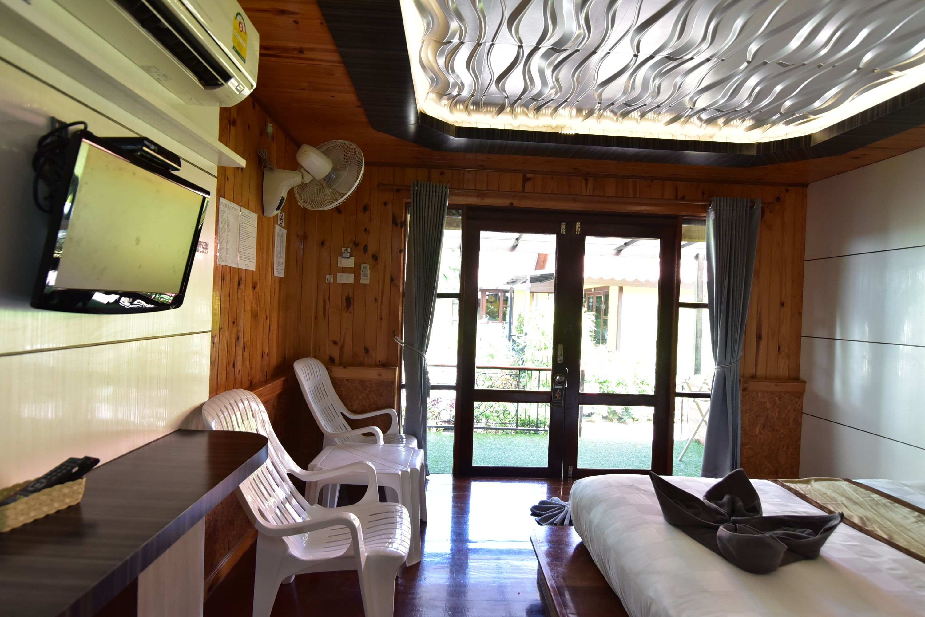 A room with a wooden ceiling, a flat-screen TV, a desk with a chair, a bed, and a view of the outside.