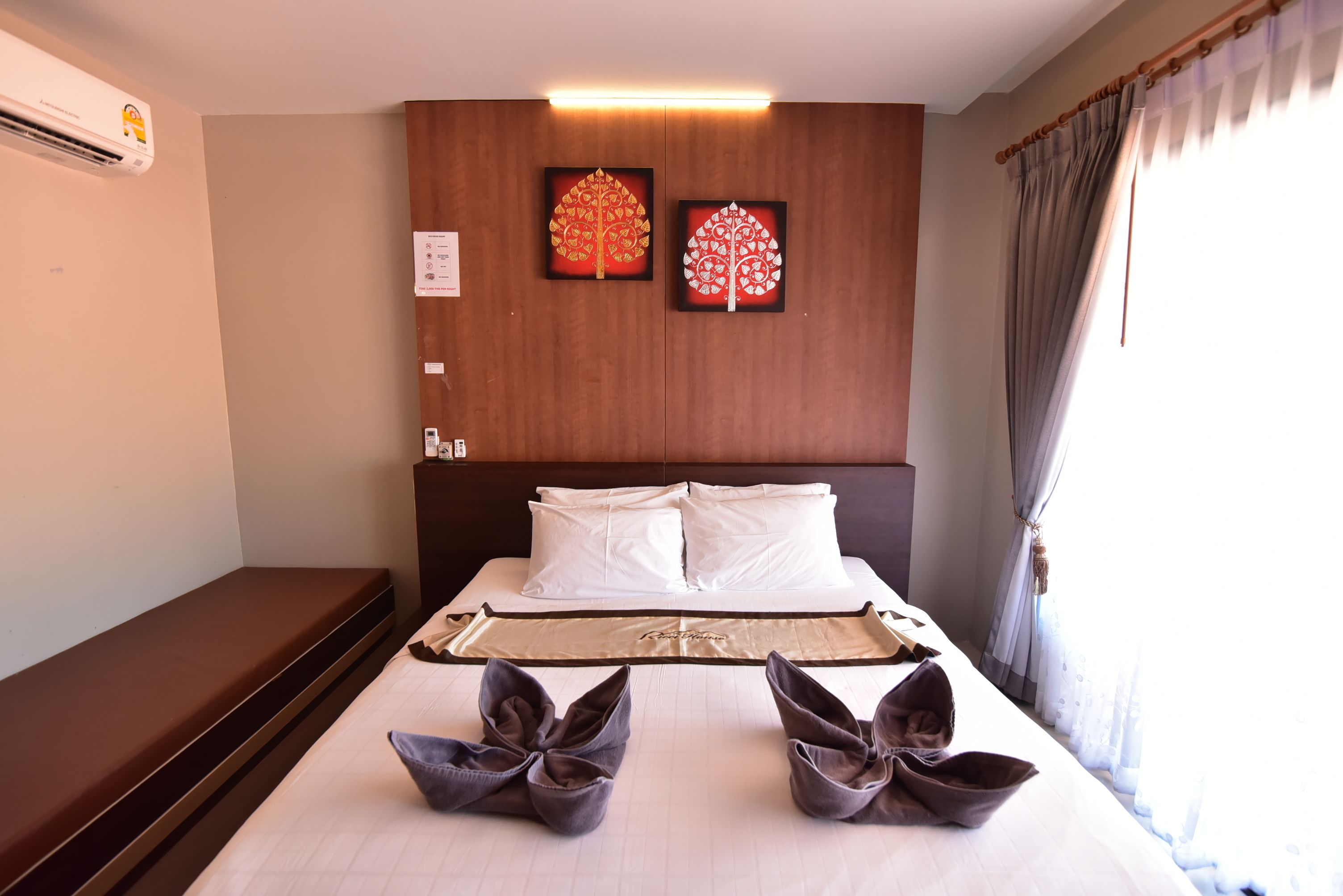 A hotel room with a bed, pillows, a headboard, artwork, a bench, and a window with curtains.