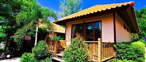 Una casita de un piso con techo de paja, rodeada de abundante vegetación y una terraza de madera.