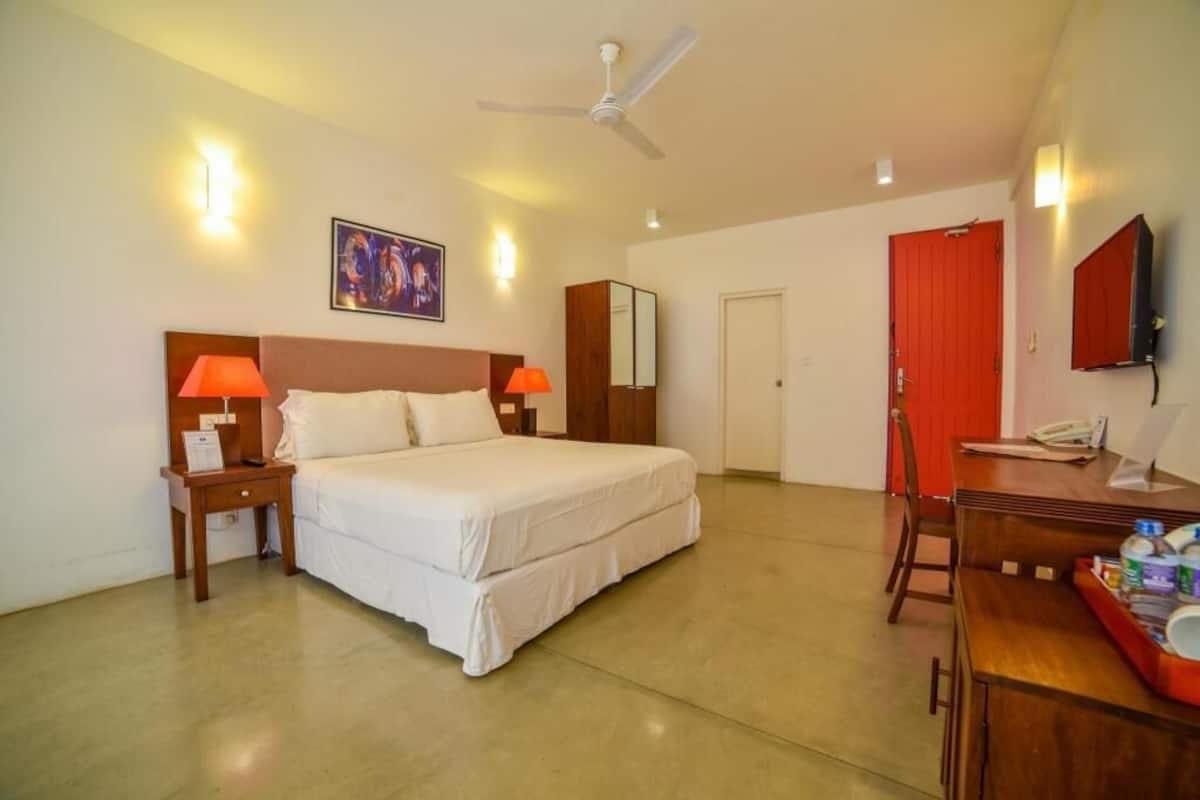 Habitación de hotel con cama, lámparas de noche, escritorio, silla, televisor, ventilador de techo y puerta.