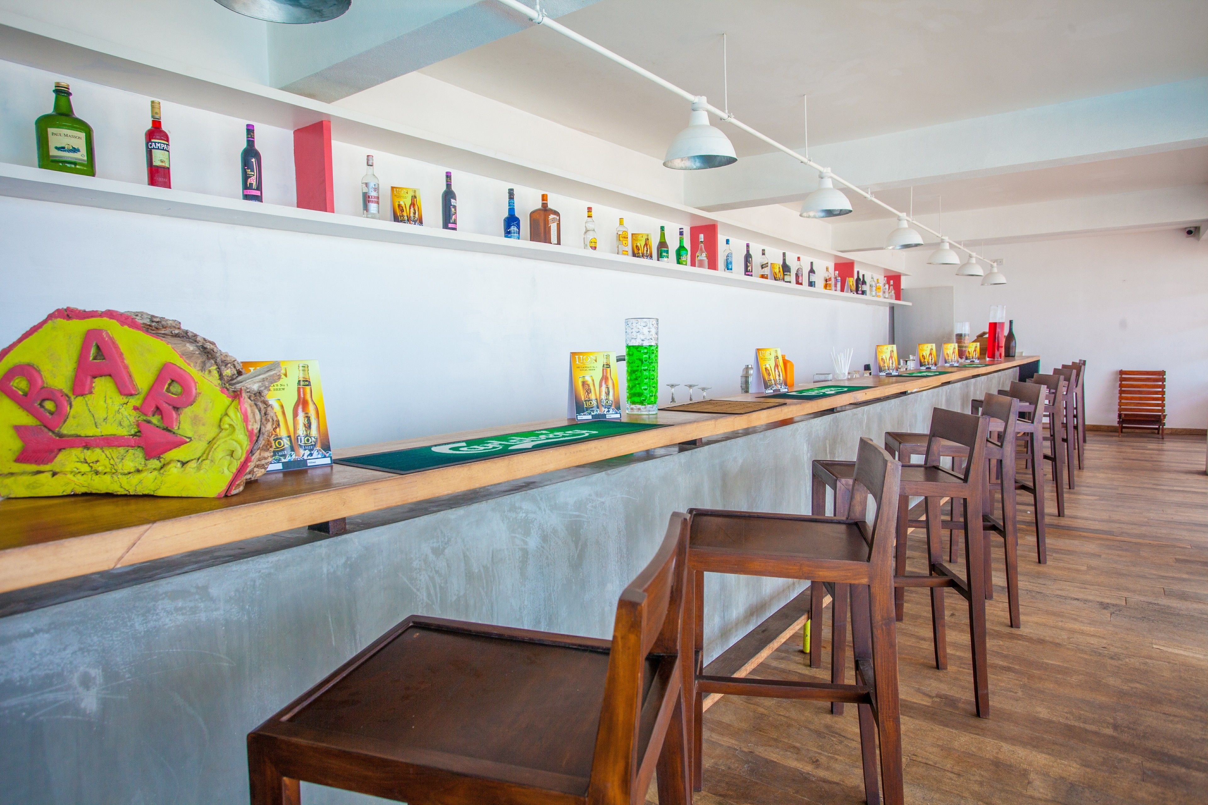Een bar met houten stoelen, een toonbank met diverse flessen en kasten vol flessen en dozen.
