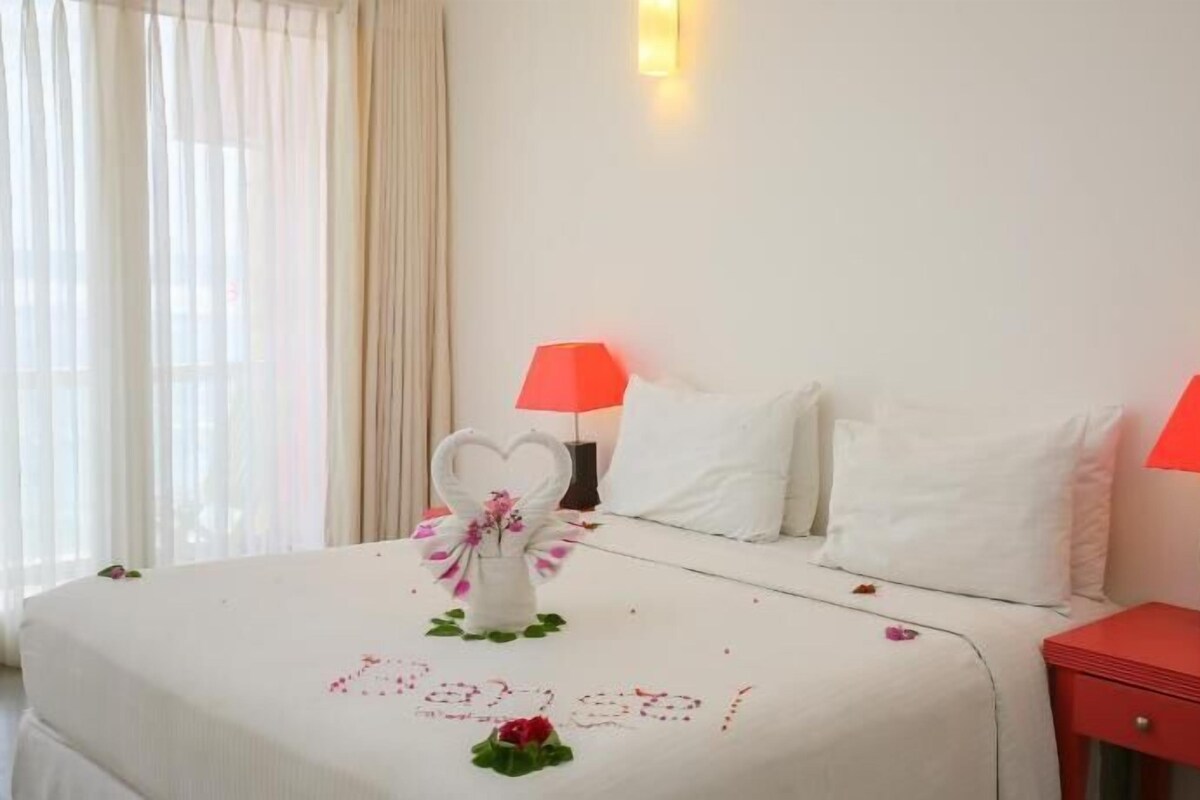 A hotel room with a bed, two bedside lamps, a window with curtains, and a decorative heart-shaped object on the bed.