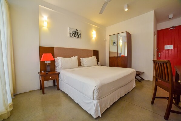 A hotel room with a large bed, a nightstand with a lamp, a wooden chair, a red door, and a ceiling fan. - Coral Rock by Bansei (Hikkaduwa)