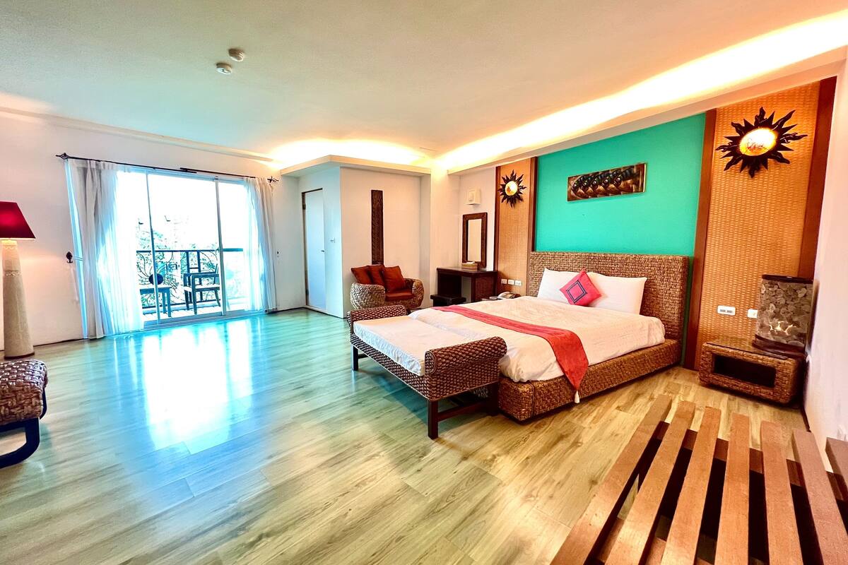 A spacious bedroom with a large bed, a wooden bench, a balcony, and a view of the outdoors.