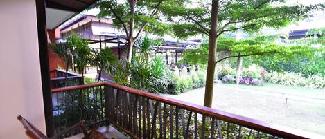 A balcony with a wooden chair, a metal railing, and a view of a garden with various plants and a building in the background.