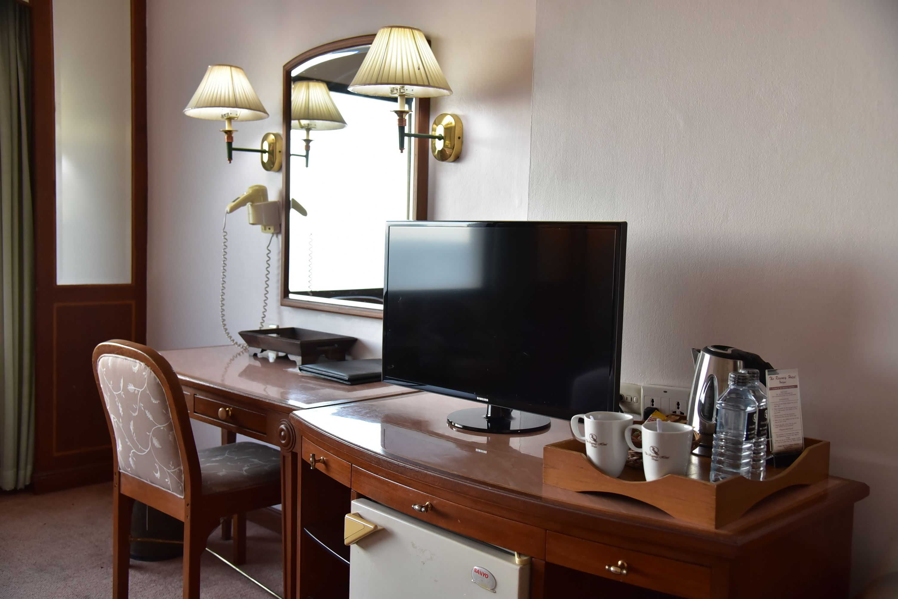 A hotel room with a wooden desk, a flat-screen TV, a desk lamp, a chair, a mini-fridge, a coffee maker, and a tray with a water bottle and a mug.
