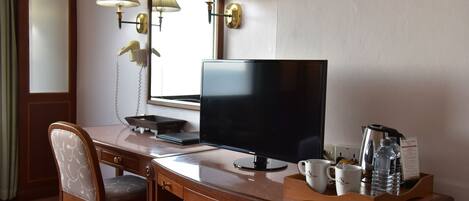 A hotel room with a wooden desk, a flat-screen TV, a desk lamp, a chair, a mini-fridge, a coffee maker, and a tray with a water bottle and a mug.