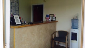 A reception area with a wooden counter, a chair, a water cooler, and a sign listing room rules. - Desert Inn (Brawley)