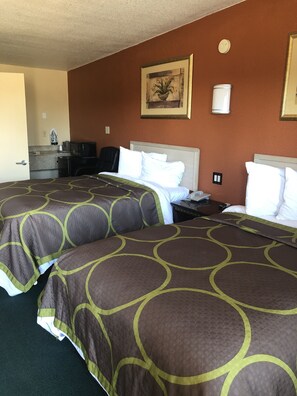 Habitación de hotel con dos camas, un escritorio, una silla, una kitchenette pequeña y un cuadro en la pared.