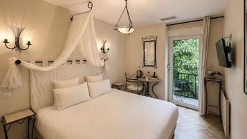 A bedroom with a bed, bedside tables, a desk, a chair, a hanging light fixture, a ceiling light, a television, and a view of greenery outside.