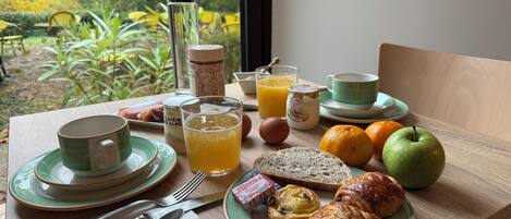 A breakfast table with plates, cups, a glass of orange juice, a jar of jam, a croissant, an apple, and a bottle of milk.