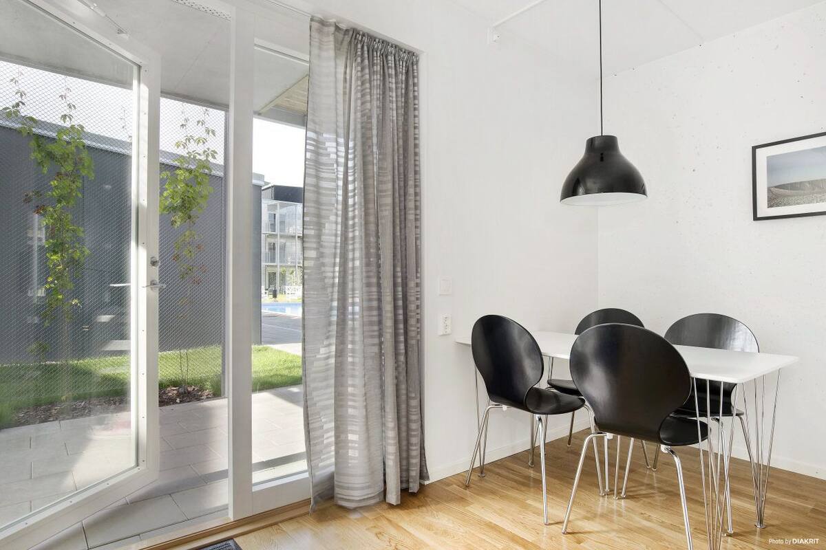 A modern dining area with a white table and black chairs, a black pendant light, and a view through a glass door to a garden.