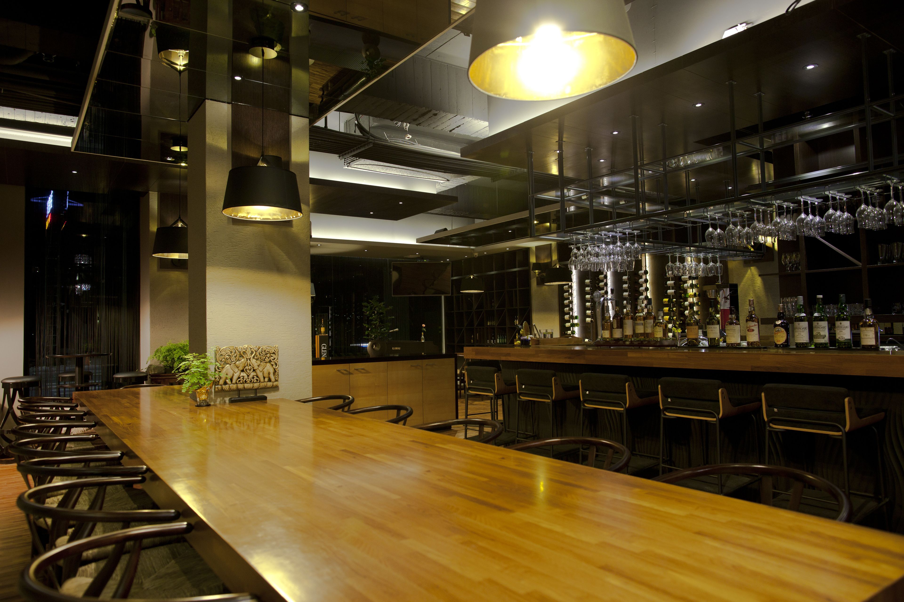 A modern bar area with a long wooden counter, multiple hanging light fixtures, and a variety of bottles on the shelves.