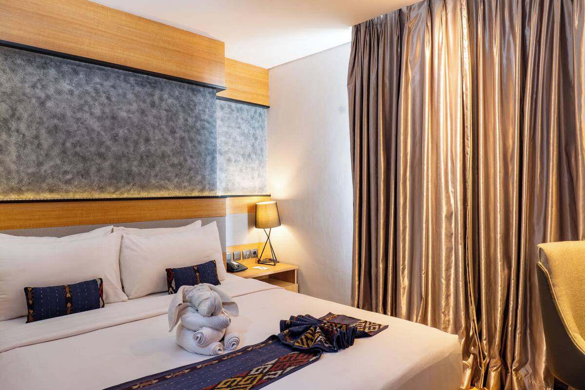 Una habitación de hotel con una cama bien hecha, cabecera, mesita de noche con lámpara y un ventanal con cortinas.