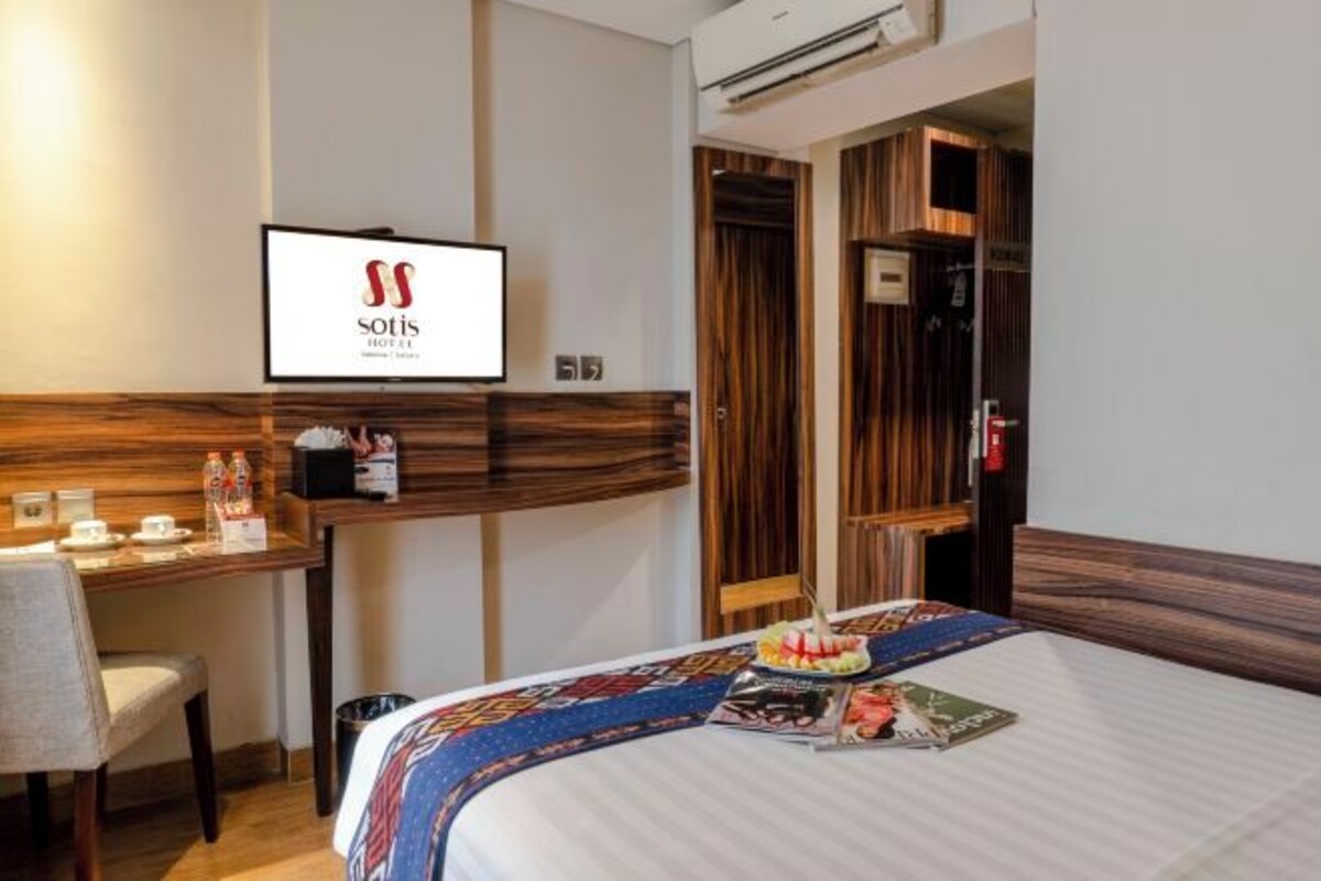 A hotel room with a bed, a desk with a chair, a TV mounted on the wall, and a wooden wardrobe.