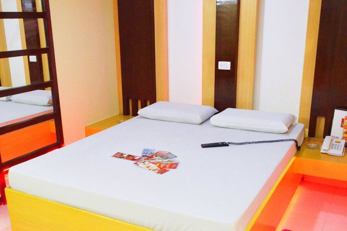 A single bed with white bedding, a bedside table with a phone and a remote control, and a mirror on the wall.