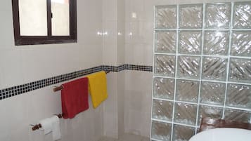Un baño con paredes de azulejos blancos, una ventana con marco marrón, un box de ducha de vidrio y dos toallas colgadas en un perchero.