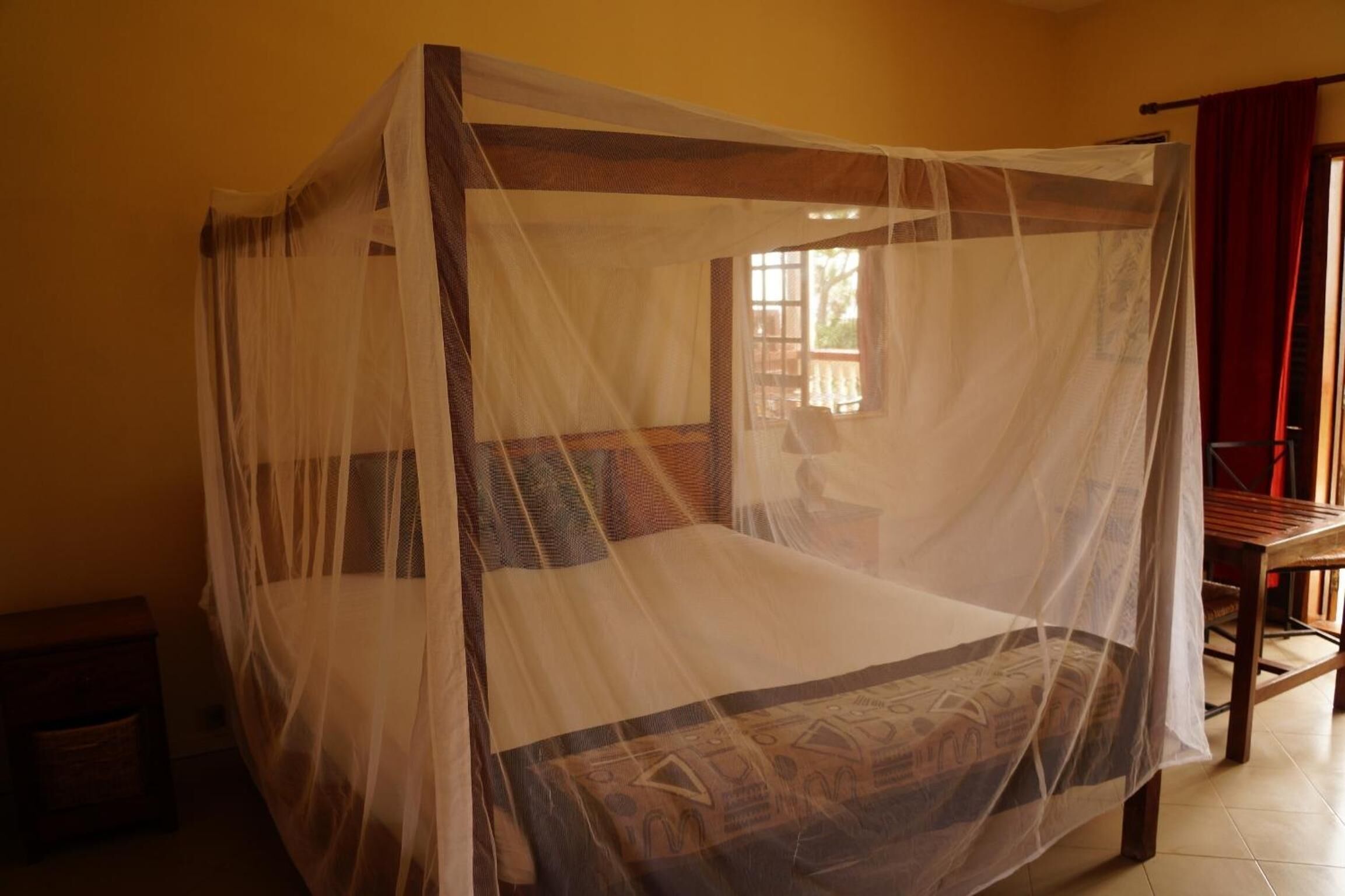 A four-poster bed with a mosquito net, a wooden nightstand, and a window with curtains.