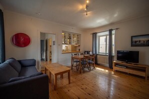 A living room with a sofa, a wooden dining table, chairs, a TV, and a red clock on the wall. - Stora Torget - Visby Lägenhetshotell  (Visby)