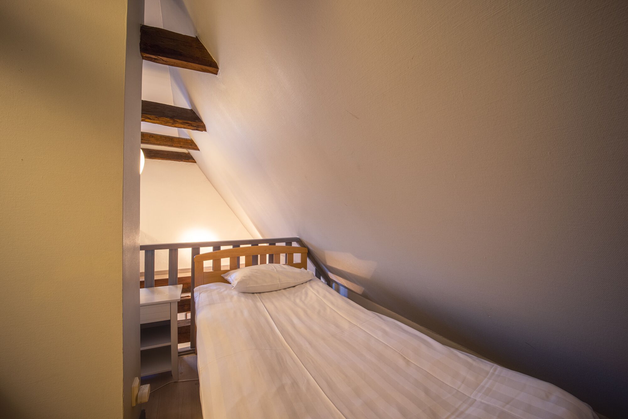 Una habitación pequeña y estrecha con techo inclinado, una cama de madera y un escritorio con silla.