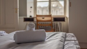 Een netjes opgemaakt bed met een gevouwen witte handdoek, een houten stoel en een raam met uitzicht op een gebouw.