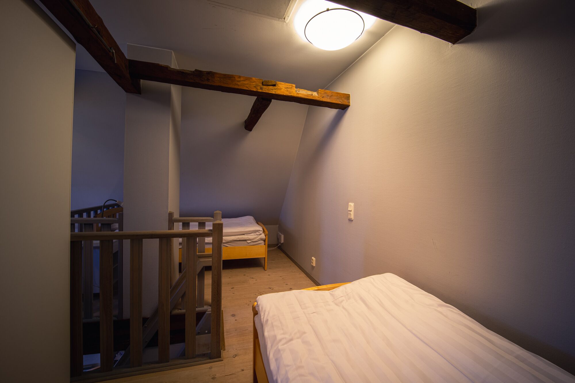 A small, dimly lit room with a sloped ceiling, a wooden bed, a small table, and a window with a view of the outside.