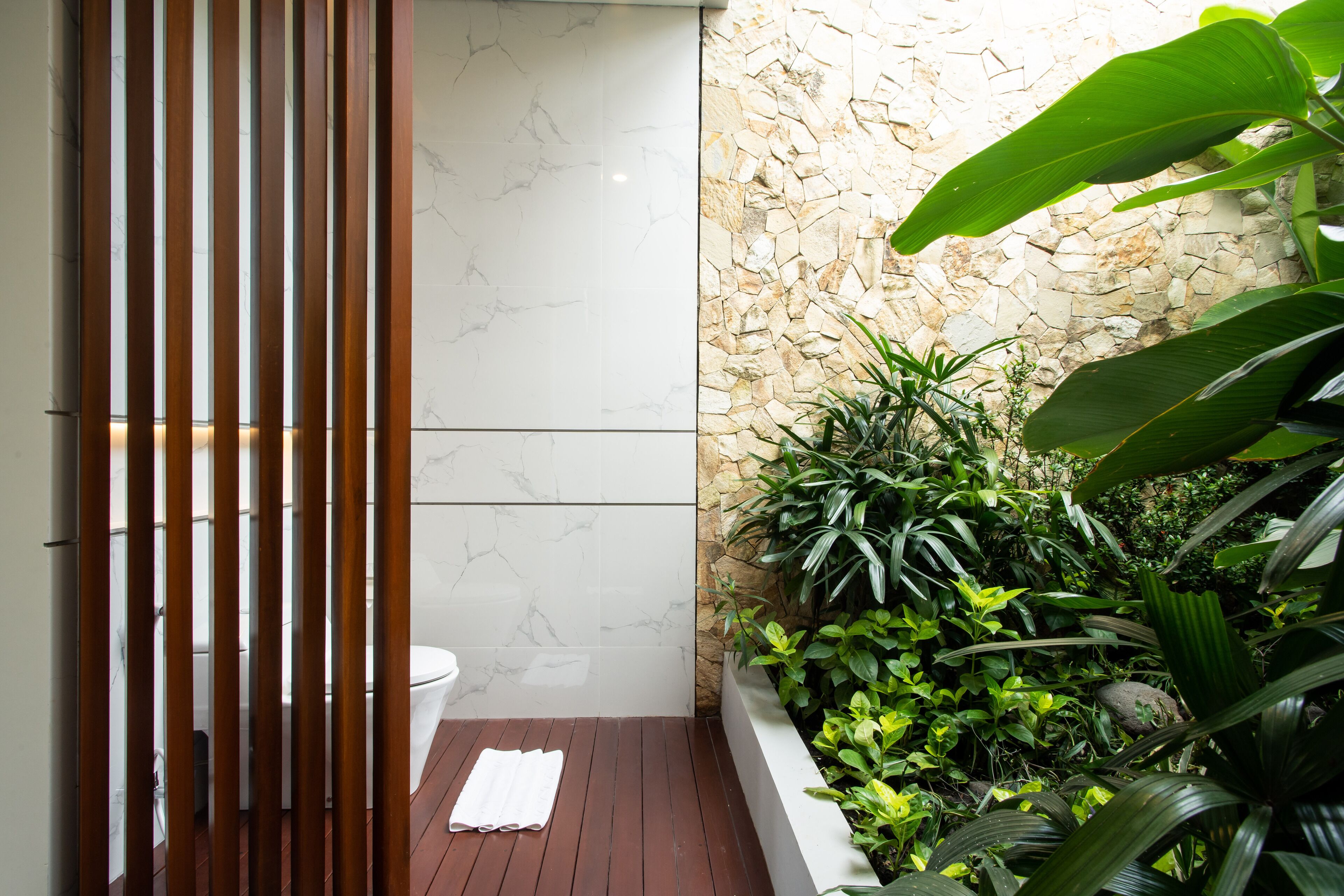 Un baño moderno con inodoro, listones de madera, pared de mármol blanco y pared de piedra con follaje.