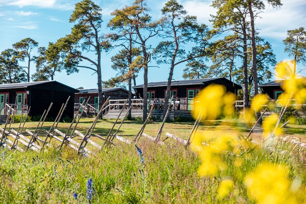 A rustic cabin with a wooden deck and a covered area, surrounded by tall trees and a grassy field. - STF Ihre Gård (Lärbro)