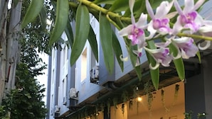 A building with a covered outdoor area, potted plants, and a white car parked in front. - At Ease Hotel Suvarnabhumi (Bang Phli)