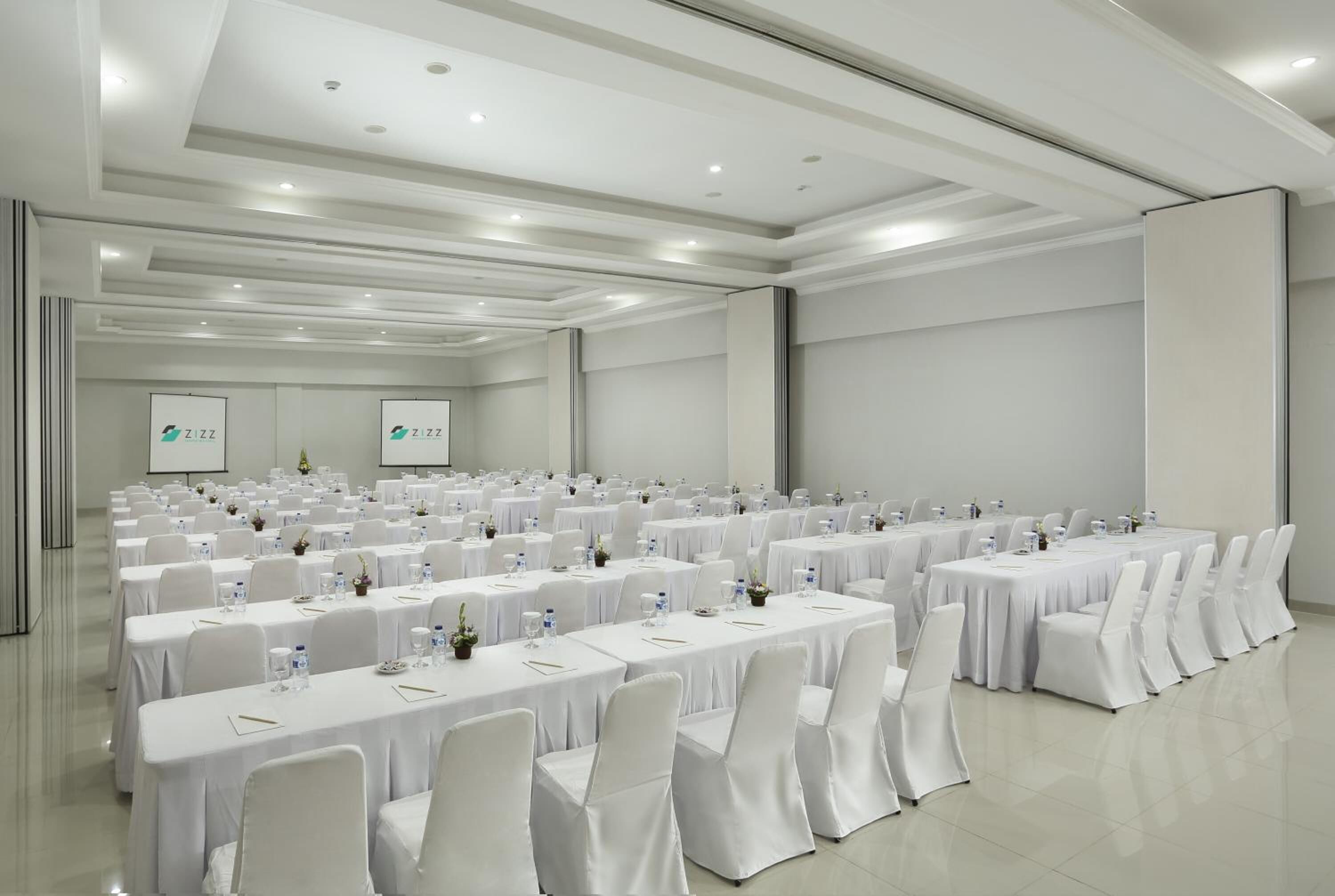 A large conference room with rows of tables and chairs, white tablecloths, and a projector screen.