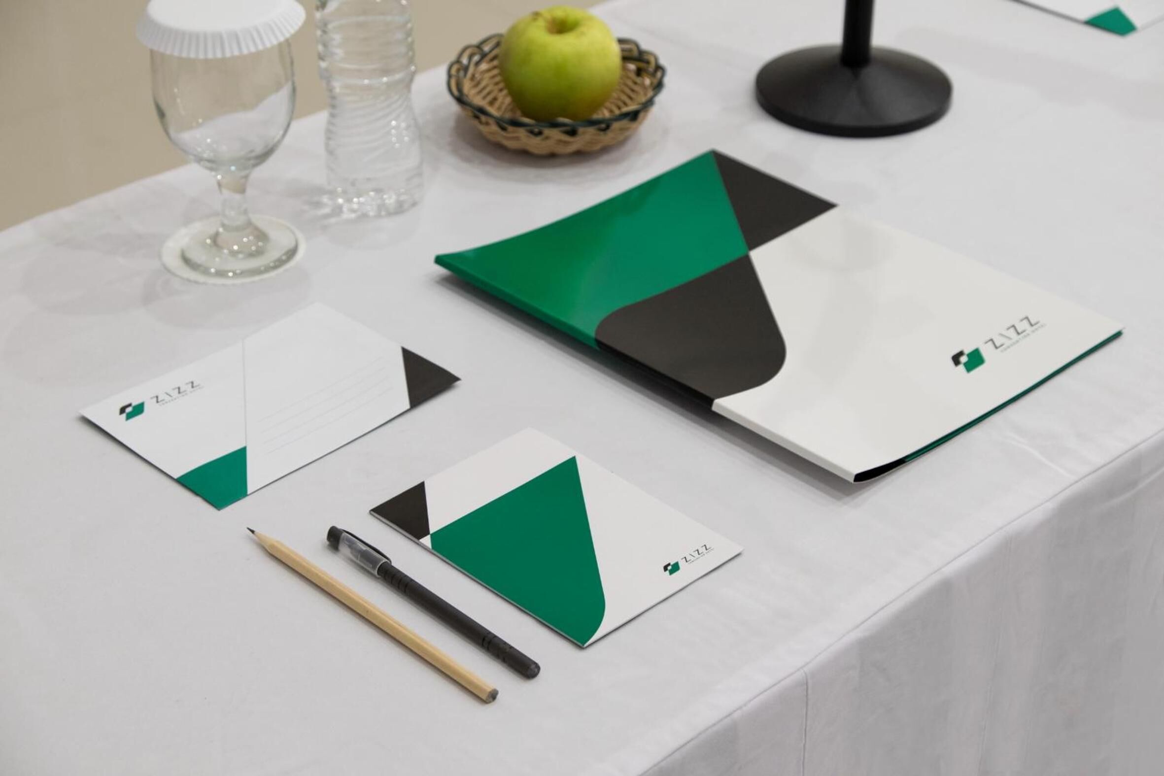 A table with a white tablecloth, a green and black folder, a pen, a glass, and a basket with an apple.