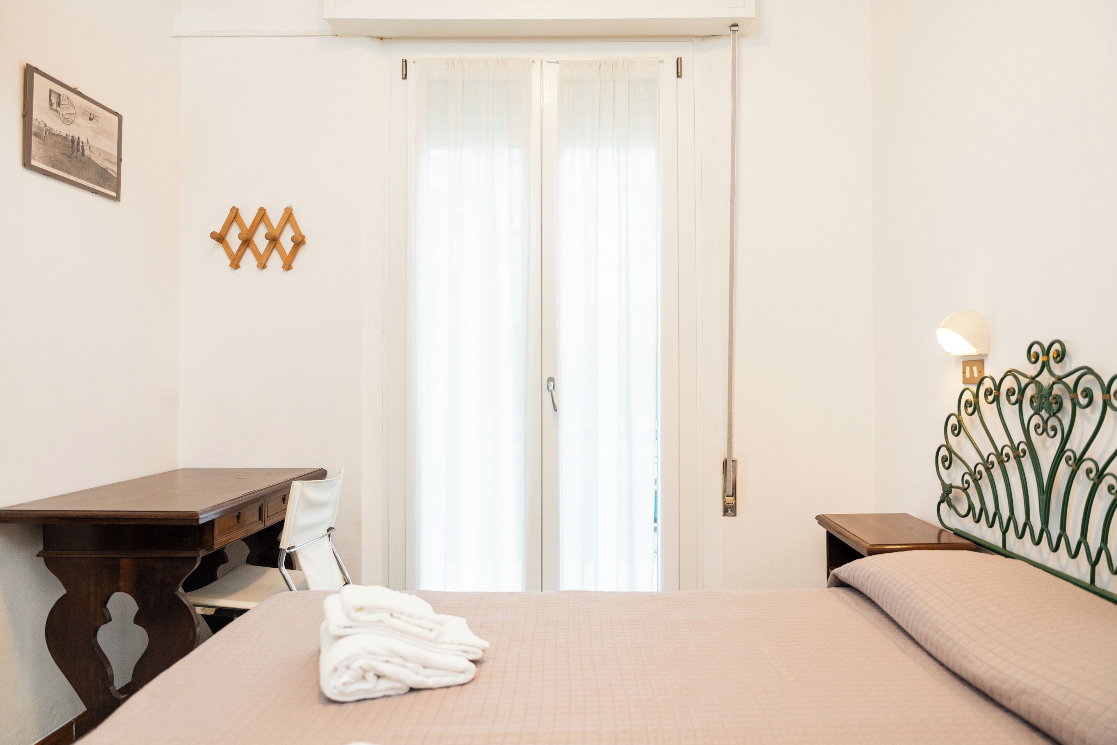 A hotel room with a bed, a desk, a chair, a nightstand, and a wall-mounted lamp.