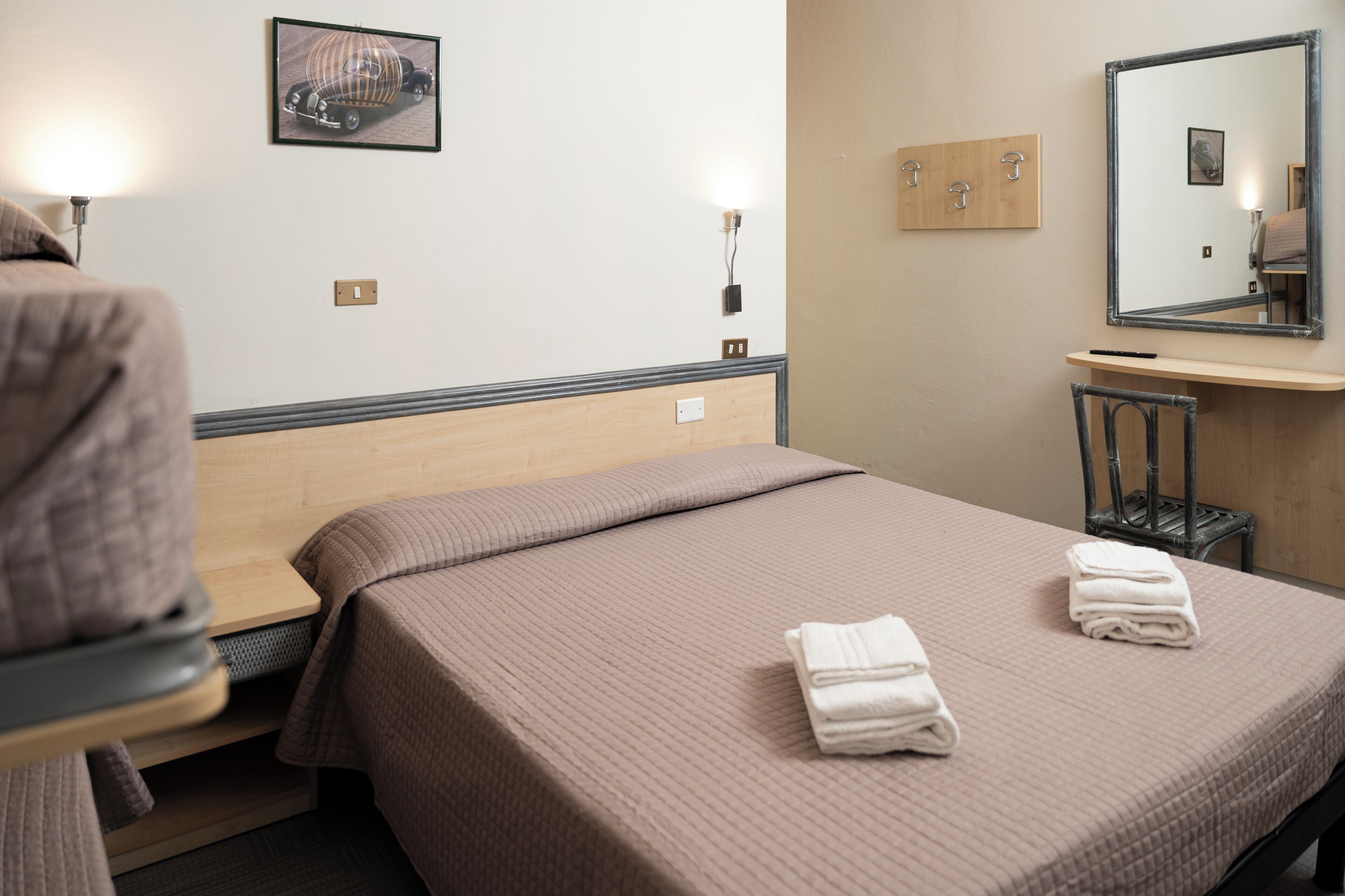 A hotel room with a bed, a chair, a desk, a mirror, and a wall-mounted picture.