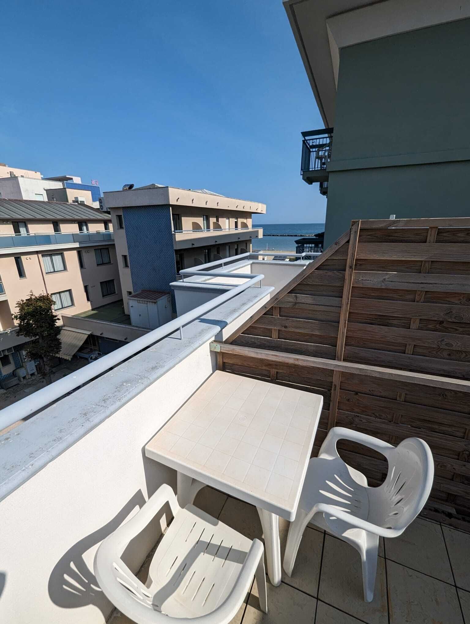 Un balcon avec une table et deux chaises, donnant sur une zone côtière avec des bâtiments et un ciel dégagé.