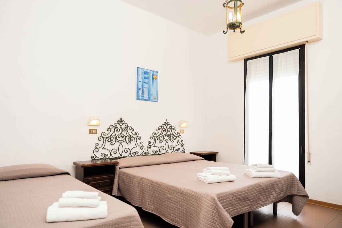 A hotel room with two beds, a decorative headboard, bedside lamps, a painting on the wall, and a sliding door to the right.