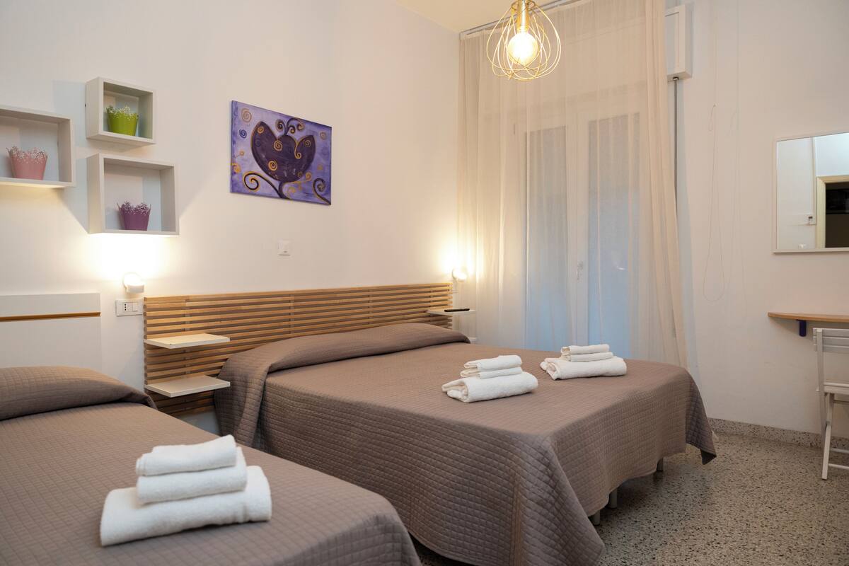 A hotel room with two beds, a wooden headboard, a painting, a window with curtains, and a bedside table with a lamp.