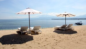 Une plage avec deux chaises longues blanches sous des parasols, un bateau sur l’eau et des montagnes en arrière-plan.
