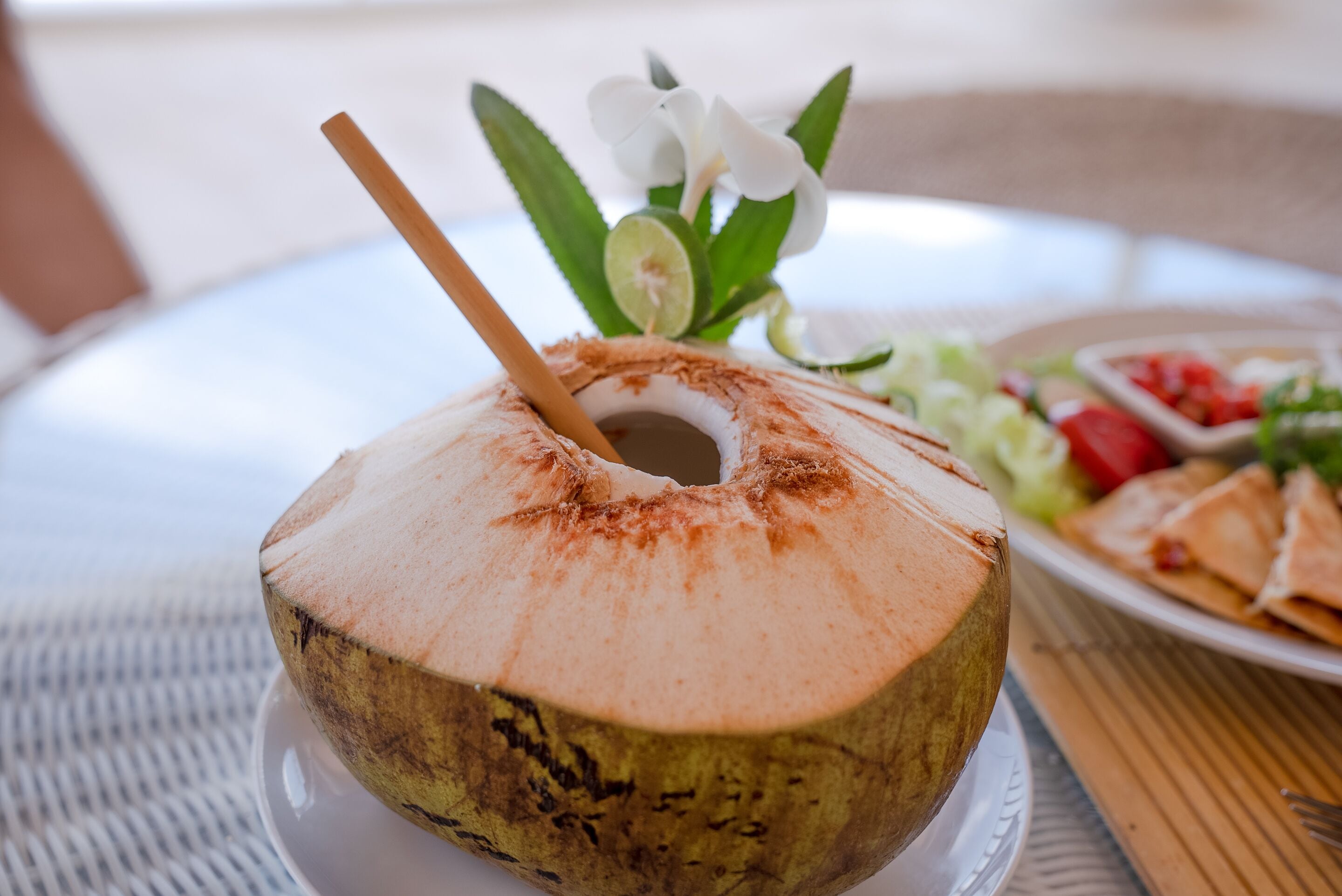 A coconut drink with a straw and a lime slice on a white plate, accompanied by a salad and a small dish of fruit.
