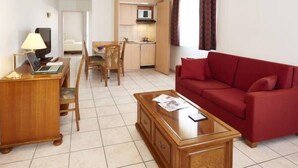 Un espacio habitable compacto con sofá rojo, mesa de centro de madera, escritorio con lámpara, televisor y una zona de kitchenette.